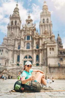 Gülümseyen kadın sırt çantalı piligrim, Camino de Santiago Yolu 'nun sonu olarak Santiago de Compostela' daki Obradeiro Meydanı 'nda (plaza) modern bir akıllı telefon kullanarak selfie çekiyor..