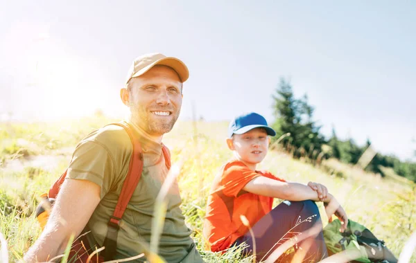 笑みを浮かべて父とティーンエイジャーの息子の肖像画に座っている草の上に彼らの取り付けハイキング中に歩く — ストック写真