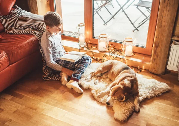 男孩躺在地板上看书 把他的小猎犬放在羊皮上 舒适的家庭环境中 温馨家庭概念形象的宁静时刻 — 图库照片