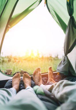 Yeşil turistik çadırın içinde çıplak ayaklı çiftin bacaklarının dikey görüntüsü. Kadın ve erkek yan yana uzanıp gün doğumunun tadını çıkarıyorlar. Aktif insanlar çadırlarla seyahat etmekten zevk alıyorlar..