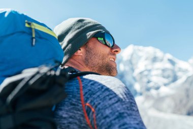 Taboche 6495 metre tepe arkaplanındaki güneş gözlüklerini koruyan sırt çantası ve ultraviyole ışınlarıyla traşlanmamış Yürüyüşçü 'nün portresi, Everest Kampı yürüyüş rotası sırasında dağ manzarasının keyfini çıkarıyor..