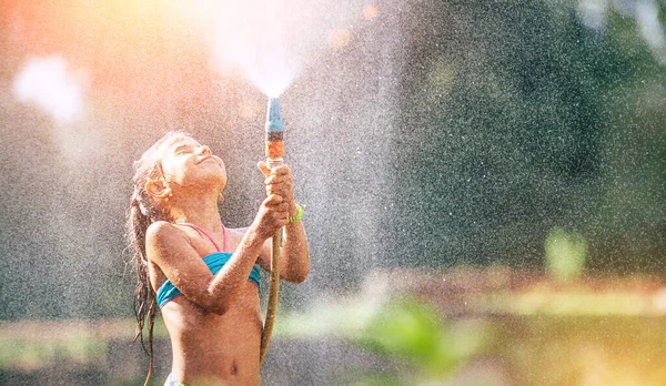 かわいい女の子がホースから自分自身を注ぐ 雨になります 暑い夏の喜び — ストック写真