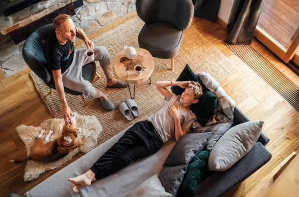 父亲和儿子一起在家里的客厅里 男孩躺在舒适的沙发上 他的爸爸抚摸着他们的小狗 对儿子笑着 和平家庭时刻概念形象 — 图库照片