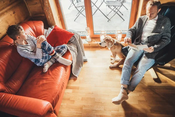 儿子和他的小猎犬在舒适的乡间小屋里度假 爸爸在看书 男孩坐着听着 狗看着窗户 — 图库照片