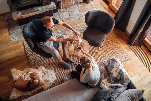 父亲和儿子一起在家里的客厅里 男孩坐在舒适的沙发上 他的爸爸把热茶倒入白杯 小猎犬躺在羊皮上 和平家庭时刻概念形象 — 图库照片