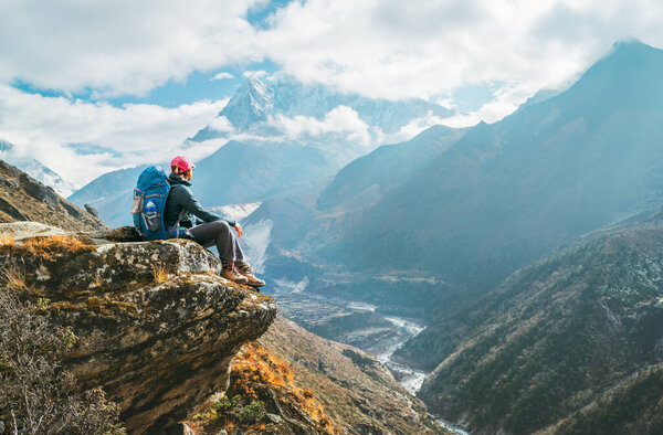 Young hiker backpacker female sitting on the cliff edge and enjoying Ama Dablam 6,812m peak view during Everest Base Camp (EBC) trekking route near Phortse, Nepal. Active vacations concept image