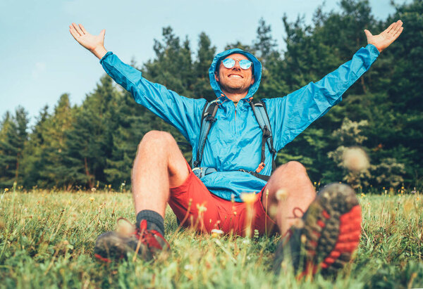Backpacker man has a rest break enjoying mountain landscape wide opened and raised arms. He wears in blue rain coat poncho and blue sunglasses. Active sports backpacking healthy lifestyle concept.