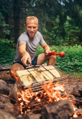 Adam kamp ateşinin üstünde ızgara ızgara ızgarasını dilimlenmiş sebzelerle tutuyor. Ormanda yaz pikniği