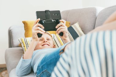Genç çocuk rahat bir kanepede uzanıyor ve şevkle oyun oynuyor akıllı telefonlu oyun alanını kullanarak.