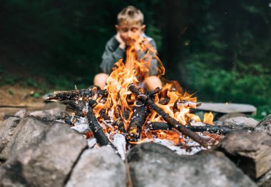 Çocuk ormanda kamp ateşinin yanında oturuyor.