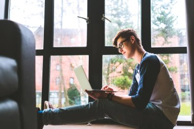 Öğrenci, pencerenin yanında duran dizüstü bilgisayarla çalışıyor.