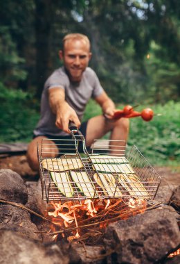 Adam ateşin üstünde, dilimlenmiş sebzelerle ızgara yapıyor. Ormanda yaz pikniği