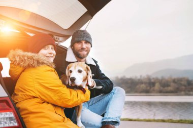Baba oğul ve av köpeği arabanın bagajında oturuyor ve kameraya gülümsüyorlar..