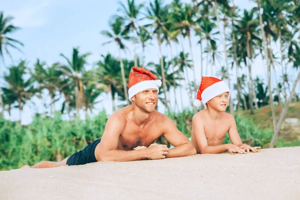 サンタ ハットの幸せな笑顔の父と息子は砂の島のビーチにサンバスを持っています — ストック写真