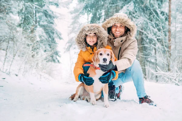 父亲和儿子穿着保暖帽衫 带着他们的小猎犬在雪地的森林里散步 笑容满面 宠物在家庭和冬季服装的概念形象 — 图库照片
