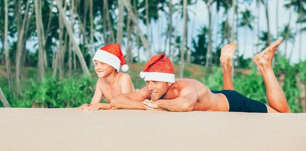 戴着圣诞礼帽快乐地微笑着的父亲和儿子在沙滩上晒日光浴 — 图库照片