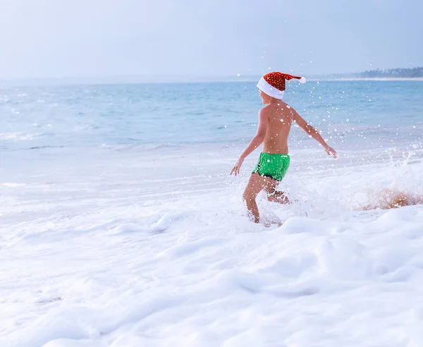 サンタの帽子の少年は海で泳ぐために走ります クリスマス休暇の時間 熱帯休暇 — ストック写真
