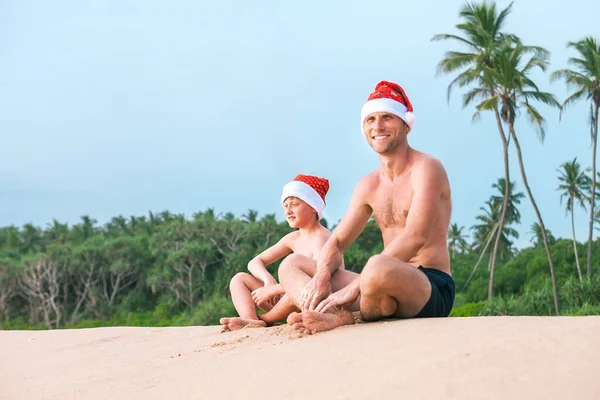 サンタの帽子の父と息子は一緒に砂の熱帯ビーチに座っています — ストック写真