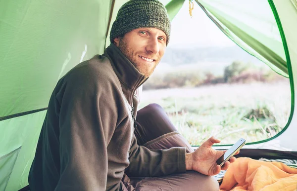 ひげを生やした旅行者暖かい服装の男はキャンプテントに座っている彼の携帯電話を使用し カメラを見て笑顔 コンセプト画像を用いた携帯電話 — ストック写真