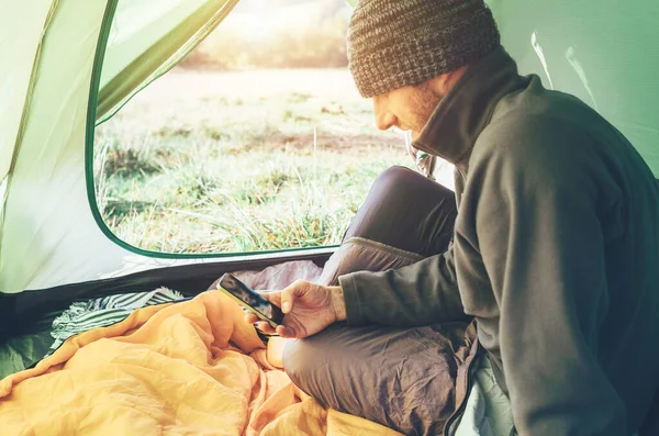 ひげを生やした旅行者暖かい服装の男はキャンプテントに座っている彼の携帯電話を使用します コンセプト画像を用いた携帯電話 — ストック写真
