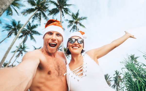 Happy smiling couple take selfie photo in Santa hats under the palm trees. New Year tropical vacation