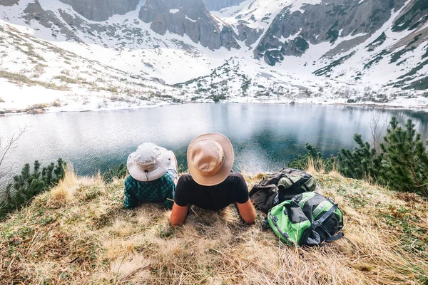 父亲和儿子的背包客们在山湖边休息 欣赏雪峰 春夏徒步旅行 — 图库照片