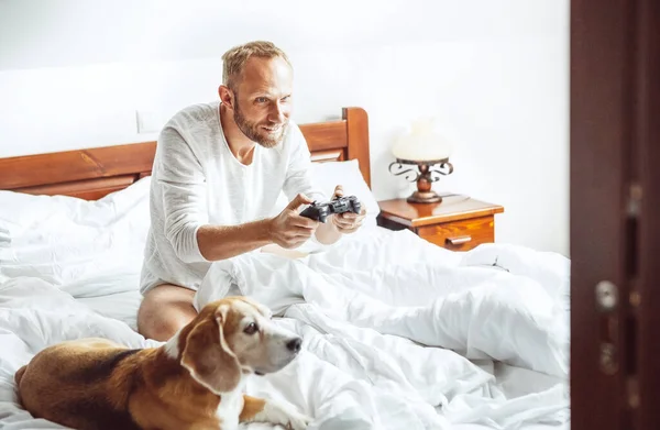 Взрослый Мужчина Проснулся Играет Компьютерные Игры Сидя Постели Собака Наблюдает — стоковое фото