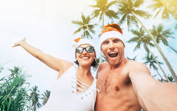 Cheerful young people couple dressed red Santa hats making selfie with tropic palm trees background Christmas image