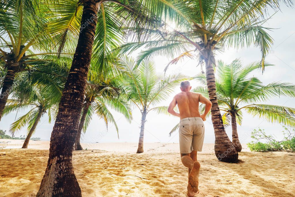Man runs under palm trees towards the ocean sand beach
