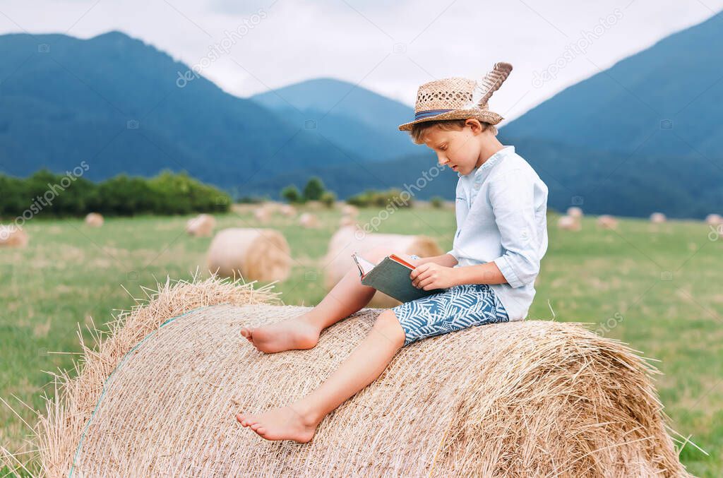 Reading boy sits over the haystack roll on the mountain field in countryside