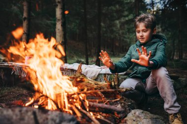 Çocuk ellerini ormanda kamp ateşinin yanında ısıtıyor.