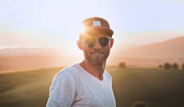 3日間の安定したひげ 野球帽 明るい夕日の太陽光線と山と牧草地の背景を持つ空想的な青いサングラスを持つカメラのハンサムな陽気な男で笑顔 多様な人々の概念 — ストック写真