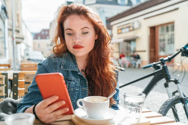 Portrait of red curled long hair caucasian teen girl sitting on a cozy cafe outdoor terrace on the street using the modern smartphone. Young woman taking a break in her city bicycle tour.