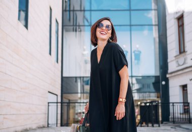 Güzel, orta yaşlı bir bayan alışveriş merkezinin yakınında siyah elbise ve güneş gözlüğü takmış kamera önünde gülüyor. Doğal güzellik insanlarının günlük konsept görüntüsü.