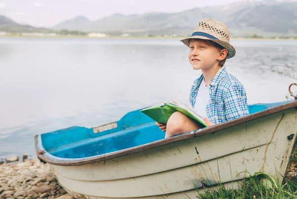 夢の少年とともに本は山の湖の銀行の古いボートに座っています — ストック写真