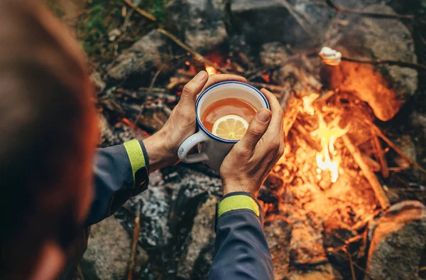 Traveler hand with big mug of hot tea near campfire