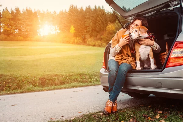 犬を持つ女性は秋の道路で車のトランクに座っています — ストック写真