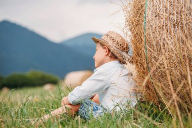 Çocuk tarlada samanlığın yanında oturuyor.