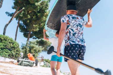 Baba sörfçülerin yanında sörfçülerle birlikte parlak güneşli bir günde raketle ayakta kürek çeken genç bir çocuk. Aktif aile yaz tatili. Deniz konsepti resminin yanında..