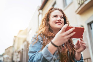 Kızıl bukleli uzun saçlı beyaz kız portresi sokakta yürüyor ve modern akıllı telefonu kullanarak internette geziniyor. Teknoloji cihazları görüntüsü olan modern insanlar.