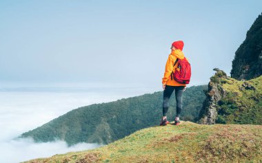 Portekiz 'in Madeira adasında, Şubat ayı sonunda bulut yolunun üstündeki dağda yürüyüş yapan turuncu su geçirmez ceket giyen genç bayan sırt çantalı. Bir dünya seyahati konsepti etrafında aktif insanlar