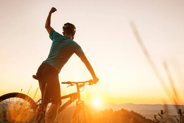 年轻人骑自行车的人在山顶碰见日落 在另一个成功的一天结束时 他举起一只手打招呼 积极运动的人的概念形象 — 图库照片