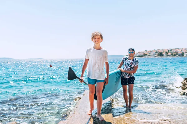 在阳光灿烂的中午 与父亲冲浪者一起 带着站着的冲浪板和划桨的金发小儿子 家庭暑期活动接近海洋概念图像 — 图库照片