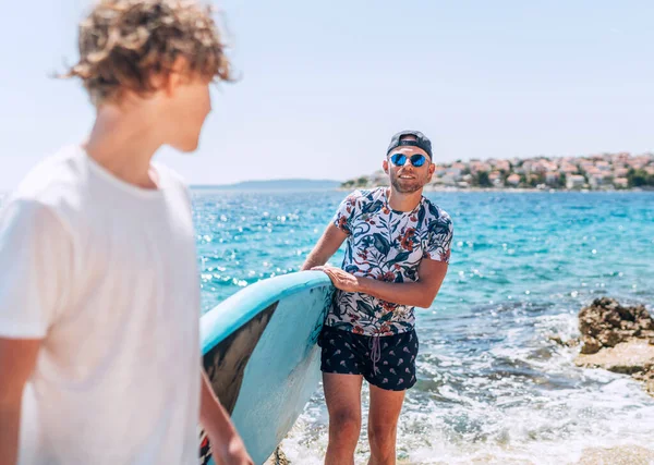 ブロンドの10代の息子は 明るい晴れた日の正午にサーフィンボードを運ぶ笑顔の父サーファーを見つめています 海のコンセプトイメージの近くでアクティブな家族の夏休み時間 — ストック写真