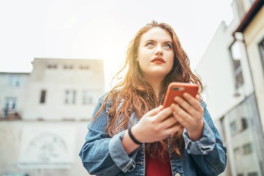 Kızıl kıvırcık saçlı beyaz kız portresi sokaklarda yürüyor ve şehir sokaklarında dolaşmak için modern akıllı telefon haritaları kullanıyor. Teknoloji cihazları görüntüsü olan modern insanlar.