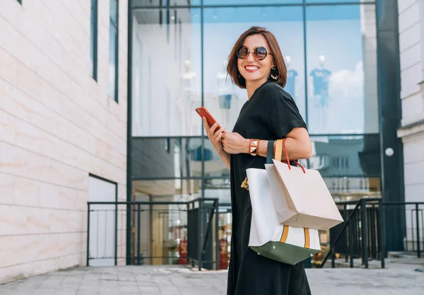 Güzel, orta yaşlı, gülümseyen, siyah elbiseli, güneş gözlüklü, elinde ince bir akıllı telefonla alışveriş merkezine giden güzel bir bayan portresi. Güzel insanlar imaj kavramını.