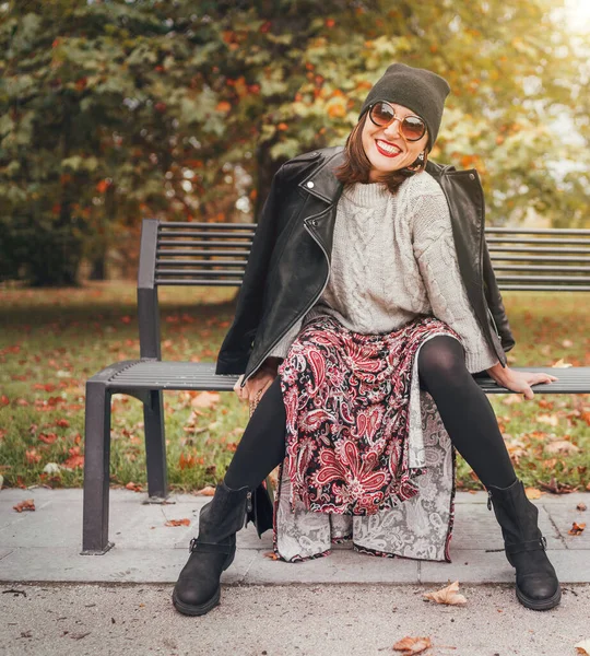 Sincerely laughing female dressed fashion boho style colorful long dress, warm knitted sweater with black leather biker jacket and Beanie Hat with flap bag sitting on bench at autumn city park