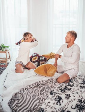 Kahkahalar atan sarı saçlı genç yetişkinler pijamalı çiftler yatak odasındaki rahat yatakta yastık savaşı yapıyorlar. Çift ilişkileri konsept resmi.