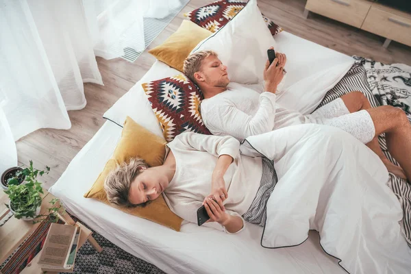 寝室の居心地の良いベッドで寝そべってインターネットを閲覧し お互いに現代的なスマートフォンを使用してメールをチェックするカップルのトップビュー 現代の技術 社会問題 関係問題の概念イメージ — ストック写真