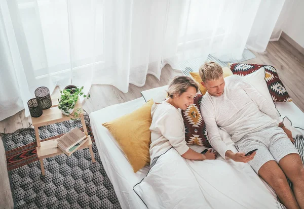 寝室の居心地の良いベッドで寝そべって インターネットを一緒に閲覧したり 現代のスマートフォンを使用してメールをチェックするカップルのトップビュー 現代の技術とカップルの関係の概念イメージ — ストック写真